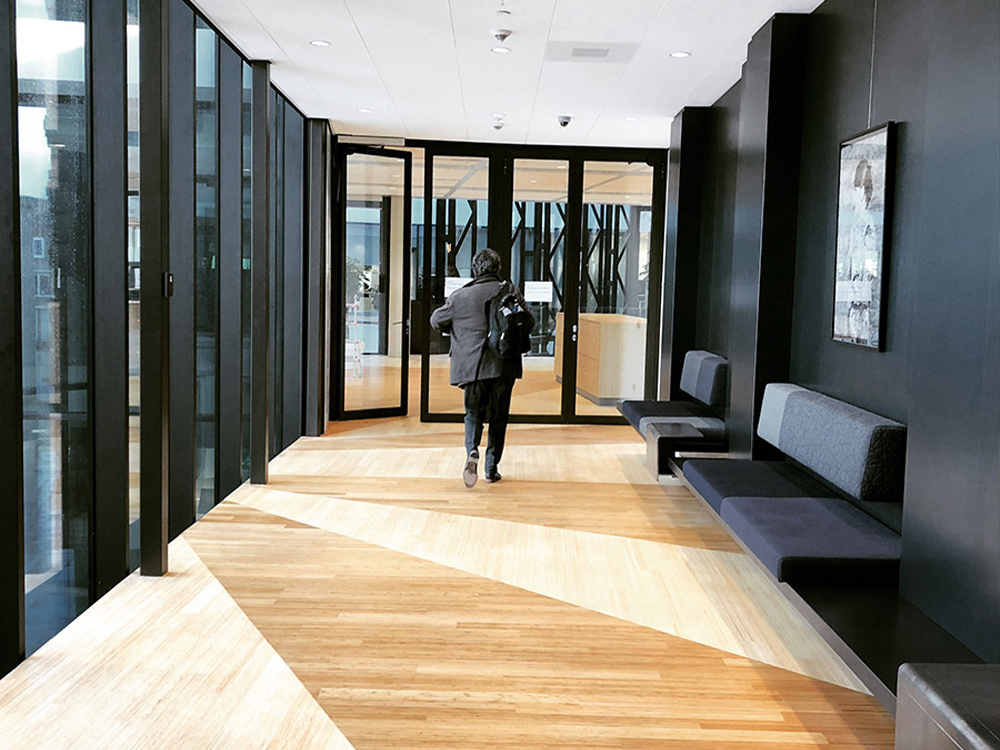 Moderne hal met houten vloer, banken en glazen deuren. Een persoon loopt naar buiten.