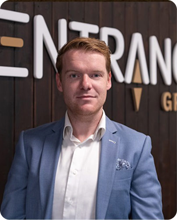 Man in een lichtblauw jasje staat voor een houten muur met 'ENTRANCE' logo.