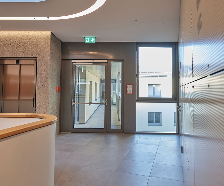 Moderne hal met lift, glazen deuren en grote ramen, lichte betegeling.