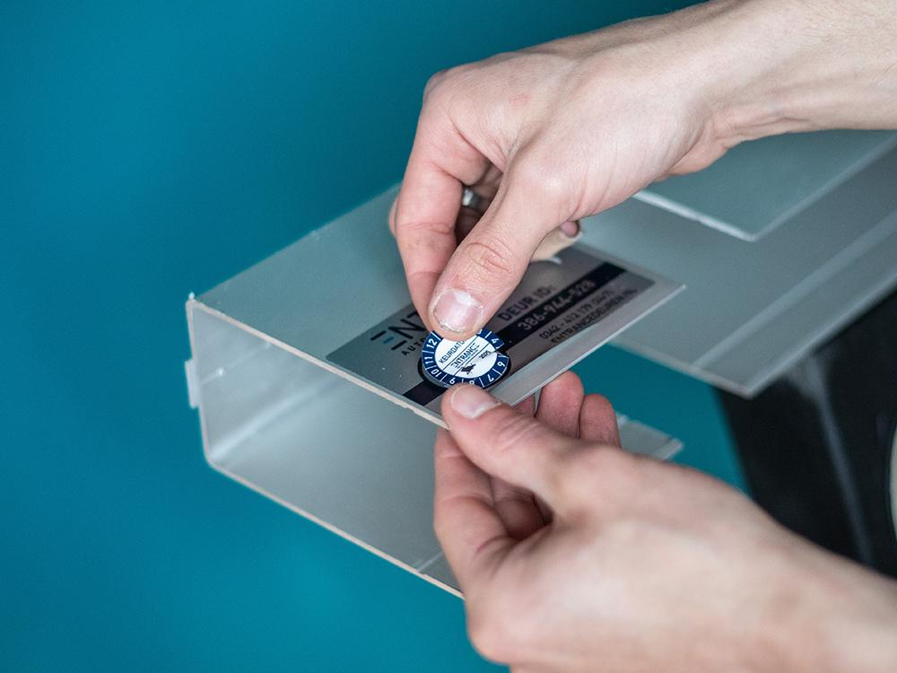 Een hand plaatst een sticker op een metalen object op een blauwe achtergrond.